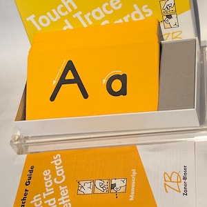 May include: A set of yellow letter cards with a teacher guide. The cards feature the letter "A" in uppercase and lowercase with arrows showing the direction to trace the letter. The guide has the text "Touch and Trace Letter Cards" and "Teacher Guide" on the front. The guide also has the text "Manuscript" and the logo for Zaner-Bloser.