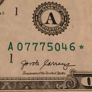 May include: Close-up of a one dollar bill with a green serial number "A 07775046*" and the signature of the Treasurer of the United States, "Jorotha Jarranz". The bill is from the Federal Reserve Bank of Boston.