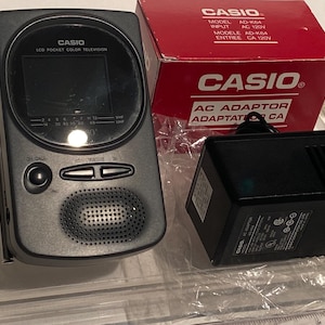 May include: A vintage Casio LCD Pocket Color Television with a black and silver finish, accompanied by a red Casio box and a black AC adaptor. The television features a small screen and speaker.