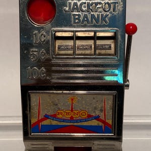 May include: A vintage silver-colored Jackpot Bank slot machine. The machine has a red handle and a red circular button. The text "JACKPOT BANK" is visible, along with the denominations 1¢, 5¢, and 10¢. The bottom of the machine features the word "RENO" and the phrase "THE BIGGEST LITTLE CITY IN THE WORLD."