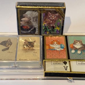 May include: A set of four decks of playing cards featuring different cat illustrations. The decks are in a clear acrylic case with a black base. The cards are labeled "Classic", "Frisk", "Congress Playing Cards", and "Congress Playing Cards".