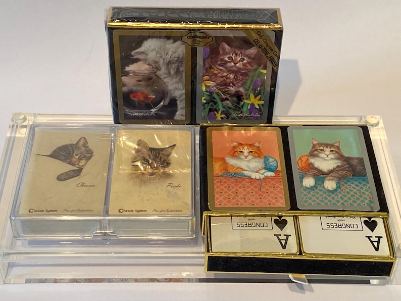 May include: A set of four decks of playing cards featuring different cat illustrations. The decks are in a clear acrylic case with a black base. The cards are labeled "Classic", "Frisk", "Congress Playing Cards", and "Congress Playing Cards".
