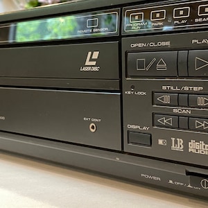 May include: A black LaserDisc player with various control buttons, including "PLAY," "OPEN/CLOSE," and "SCAN." The player has the "LASER DISC" logo and "LB digital AUDIO" text. The model number LD-V8000 is visible.