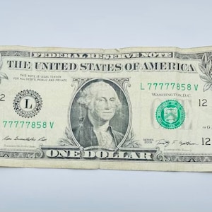 May include: A one dollar bill featuring a portrait of George Washington. The bill is green and white with the text "THE UNITED STATES OF AMERICA" and "ONE DOLLAR" printed on it. The serial number is L77777858V.