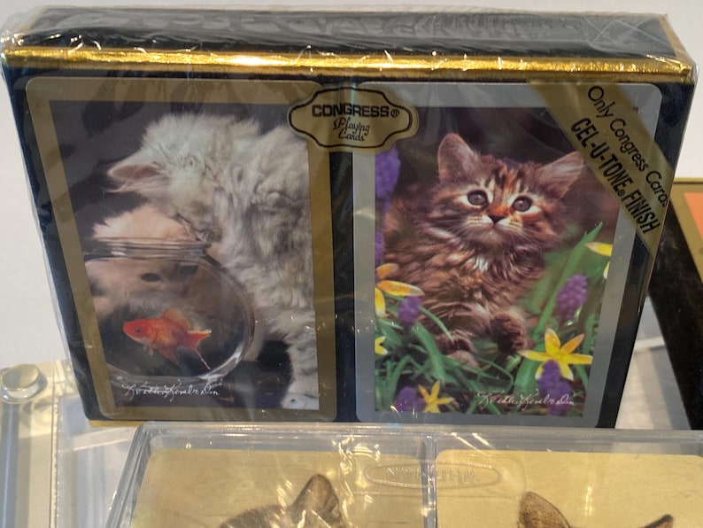 May include: A deck of playing cards with a white cat in a fishbowl on one side and a brown tabby cat in a field of flowers on the other. The cards are from Congress Playing Cards and have a Gel-U-Tone finish. The text "Only Congress Cards Gel-U-Tone Finish" is printed on the back of the cards.