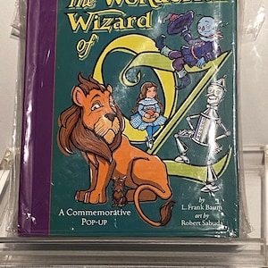 May include: A commemorative pop-up book titled "The Wonderful Wizard of Oz." The cover features illustrations of the Cowardly Lion, Dorothy, the Scarecrow, the Tin Man, and the Wicked Witch of the West. The book is sealed in plastic.