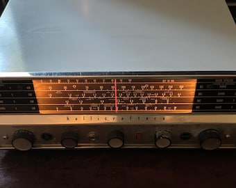 May include: A vintage Hallicrafters radio receiver featuring a metallic silver casing and a horizontal illuminated display panel with black frequency markings. The front control panel includes five black circular knobs, a headphone jack, and a red indicator needle.