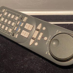 May include: A gray JVC remote control with a circular jog shuttle button. The remote has buttons for power, cancel, search, enter, OSD, memory, select, volume, channel, menu, and a forward and reverse button.