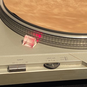 May include: Silver turntable with a wooden platter. Features a speed selector for 33 and 45 RPM records, and a pitch adjustment knob. A clear, pink, prism-shaped object sits on the turntable's base.