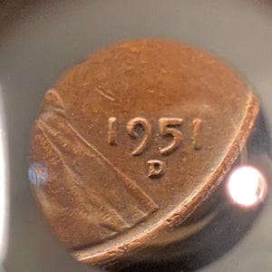 May include: Close-up of a copper-colored coin. The coin features the year "1951" and the letter "D". The coin has a worn appearance, with visible details of the design.