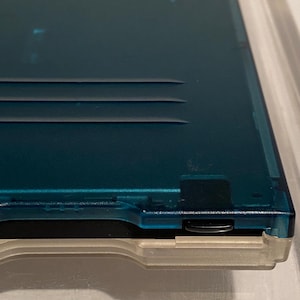 May include: A teal-colored, translucent plastic case with a light gray base. The case has three horizontal grooves and a small black button on the side. The design suggests it is a storage case for a small electronic device.