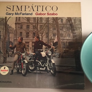 Może przedstawiać: Okładka winylowego albumu z epoki przedstawiająca dwóch mężczyzn na motocyklach w miejskim otoczeniu. Tytuł albumu to "SIMPÁTICO" z wymienionymi poniżej artystami: "Gary McFarland" i "Gabor Szabo". Album jest nagrany w mono na wytwórni Impulse!.