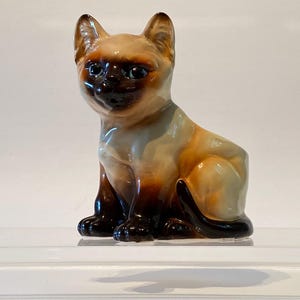 May include: A ceramic figurine of a Siamese cat sitting with its tail curled around its legs. The cat is light brown and cream with dark brown markings on its face and legs.