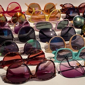 May include: A collection of sunglasses in various colors and styles. The sunglasses include shades of pink, orange, yellow, purple, green, and brown. Frames vary from round to square, with both dark and light-colored frames.