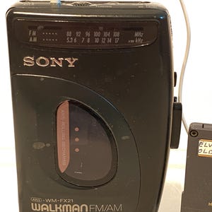 May include: A black Sony Walkman FM/AM radio with a white earphone cable plugged in. The radio has a tuning dial and frequency display. The text "WALKMAN FM/AM" is visible on the front.