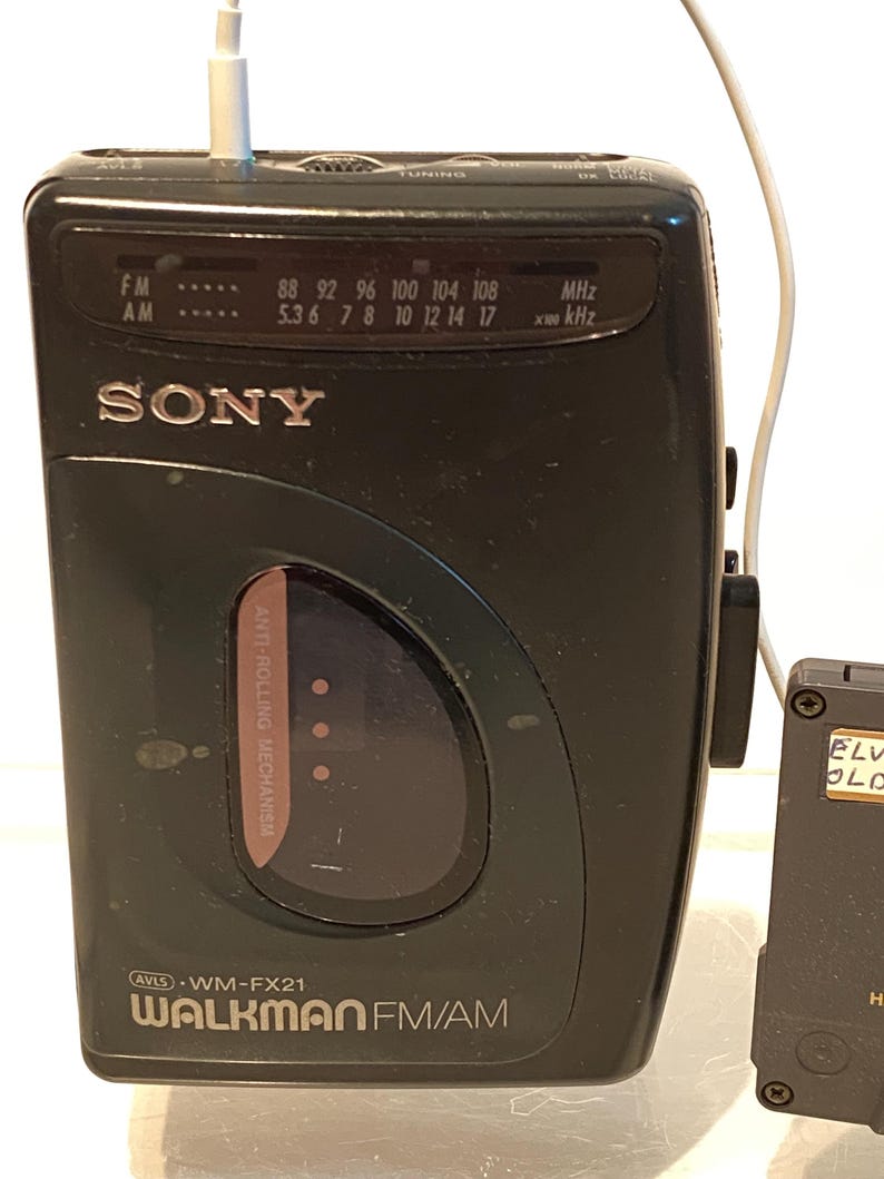 May include: A black Sony Walkman FM/AM radio with a white earphone cable plugged in. The radio has a tuning dial and frequency display. The text "WALKMAN FM/AM" is visible on the front.