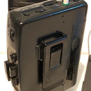 May include: Black Sony Walkman cassette player with a silver headphone jack. The device has buttons, a volume dial, and a belt clip. The word "SONY" is visible on the back.