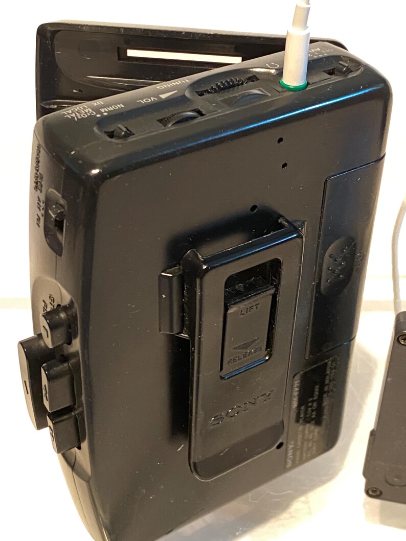 May include: Black Sony Walkman cassette player with a silver headphone jack. The device has buttons, a volume dial, and a belt clip. The word "SONY" is visible on the back.