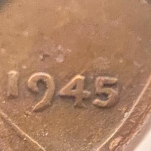 May include: Close-up of a copper-colored coin with the year 1945 prominently displayed. The numbers are embossed and slightly worn, set against a textured background. The coin's edges are partially visible, suggesting a circular shape.