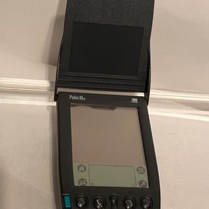 May include: A black Palm IIIe personal digital assistant (PDA) with a flip-up cover. The screen displays a simple interface with icons. The device has several buttons and a green power button. The model name "Palm IIIe" is printed on the device.