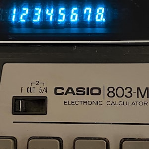 May include: A vintage Casio 803-M electronic calculator with a blue digital display showing the numbers 12345678. The calculator has a gray plastic body with black buttons labeled "R", "M-", "M+", and "÷".