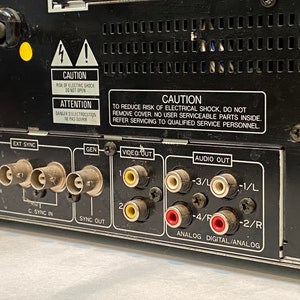 May include: Close-up of the back panel of a black electronic device. It features various input and output ports, including EXT SYNC, GEN, VIDEO OUT, and AUDIO OUT. Warning labels are visible, with text in English and French.