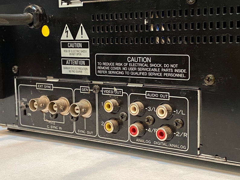 May include: Close-up of the back panel of a black electronic device. It features various input and output ports, including EXT SYNC, GEN, VIDEO OUT, and AUDIO OUT. Warning labels are visible, with text in English and French.