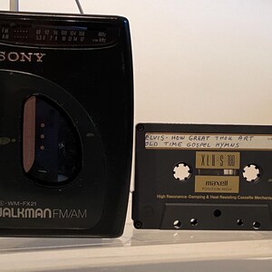 May include: A black Sony Walkman FM/AM radio and a black cassette tape. The Walkman has the text "SONY WALKMAN FM/AM" and a frequency dial. The cassette tape reads "ELVIS - HOW GREAT THOU ART OLD TIME GOSPEL HYMNS" and "XLD S 100".