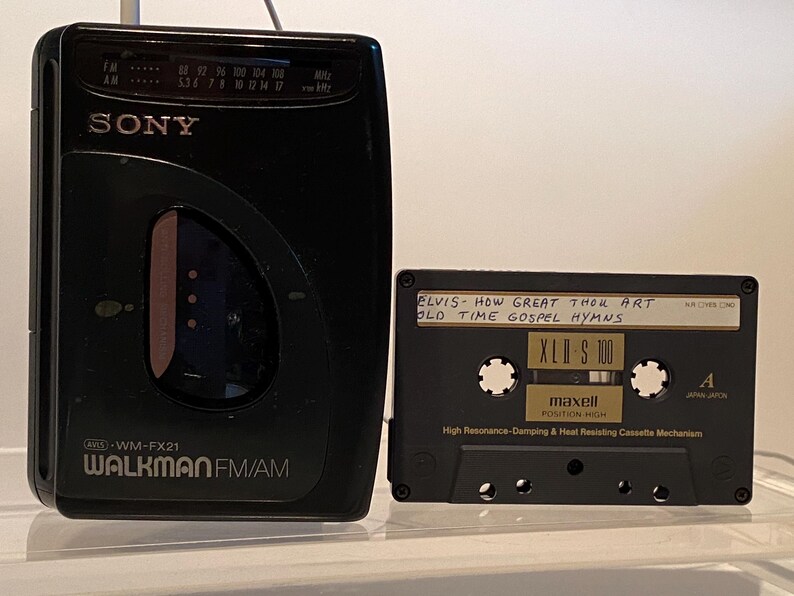 May include: A black Sony Walkman FM/AM radio and a black cassette tape. The Walkman has the text "SONY WALKMAN FM/AM" and a frequency dial. The cassette tape reads "ELVIS - HOW GREAT THOU ART OLD TIME GOSPEL HYMNS" and "XLD S 100".