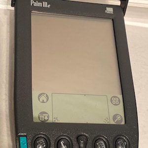 May include: A black Palm IIIe handheld personal digital assistant (PDA) with a flip-up cover. The device has a monochrome screen and several buttons below the screen. The text "Palm IIIe" is visible on the top of the device.