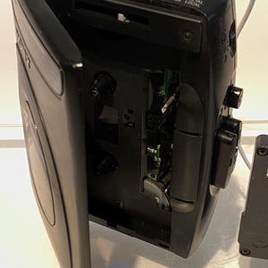 May include: Black vintage Sony Walkman cassette player with the cassette door open. The player has buttons for tuning, volume, and other functions. A white cable and green jack are plugged into the top. The player is on a white surface.