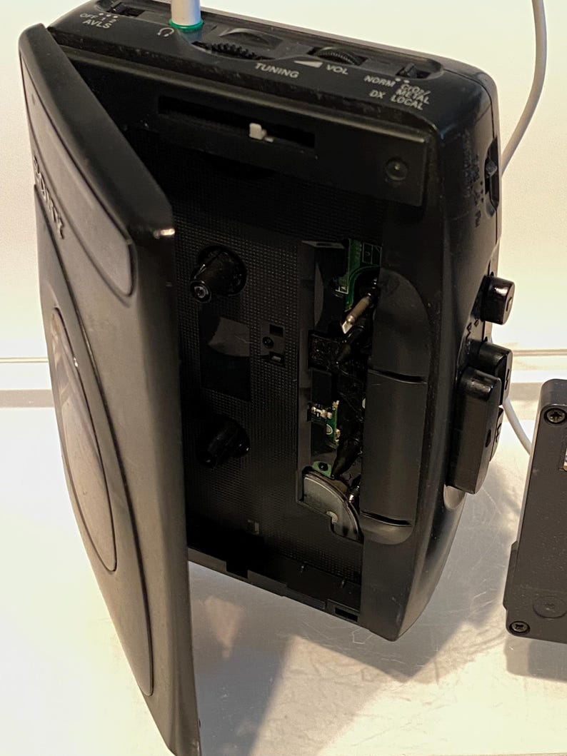 May include: Black vintage Sony Walkman cassette player with the cassette door open. The player has buttons for tuning, volume, and other functions. A white cable and green jack are plugged into the top. The player is on a white surface.