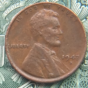 May include: A copper-colored 1947 US penny coin with the words "IN GOD WE TRUST" and "LIBERTY" inscribed on it. The coin features a profile of Abraham Lincoln on the obverse side.