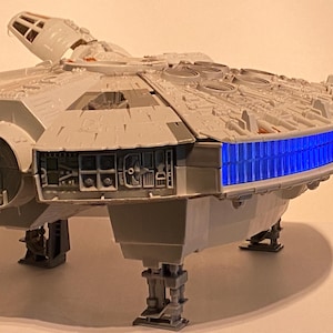 May include: A detailed model of the Millennium Falcon spaceship, featuring a light gray exterior with intricate paneling and a blue illuminated section. The model is elevated on landing gear, showcasing its iconic design.