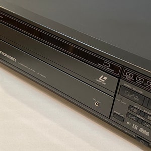 May include: A dark gray Pioneer LaserDisc player. The player has several buttons and a display panel. The text "LaserDisc" and "LB digital audio" are visible. The model number is LV-V8000G.