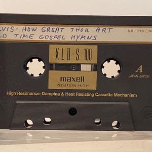 May include: A black Maxell XLII-S 100 audio cassette tape with handwritten text reading "ELVIS - HOW GREAT THOU ART OLD TIME GOSPEL HYMNS." The cassette has a gold label and the text "POSITION-HIGH".