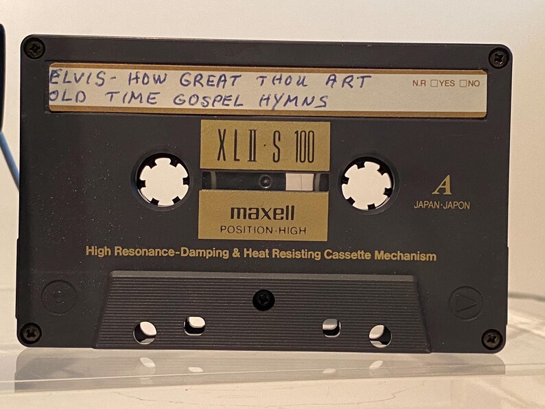 May include: A black Maxell XLII-S 100 audio cassette tape with handwritten text reading "ELVIS - HOW GREAT THOU ART OLD TIME GOSPEL HYMNS." The cassette has a gold label and the text "POSITION-HIGH".