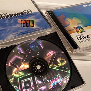 May include: Three Microsoft software CD-ROMs. The top two cases are labeled "Windows 98 Second Edition Updates" and "Office XP Professional." The bottom CD is in an open black case. The CDs have colorful, reflective surfaces.