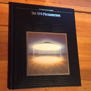 May include: A black book with a silver title "Mysteries of the Unknown: The UFO Phenomenon". The cover features a black and white illustration of a flying saucer over a desert landscape.