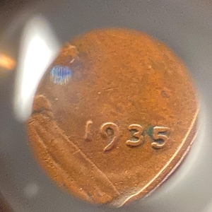 May include: A close-up of a copper-colored coin with the year 1935 visible. The coin has some wear and tear, with a slightly damaged edge. The coin is likely a vintage penny.