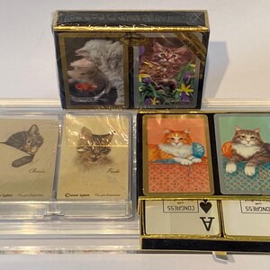 May include: A set of four playing card decks featuring different cat illustrations. The decks are in a clear plastic case with a black base. The front of the case has the text "CONGRESS" and "A" for Ace.