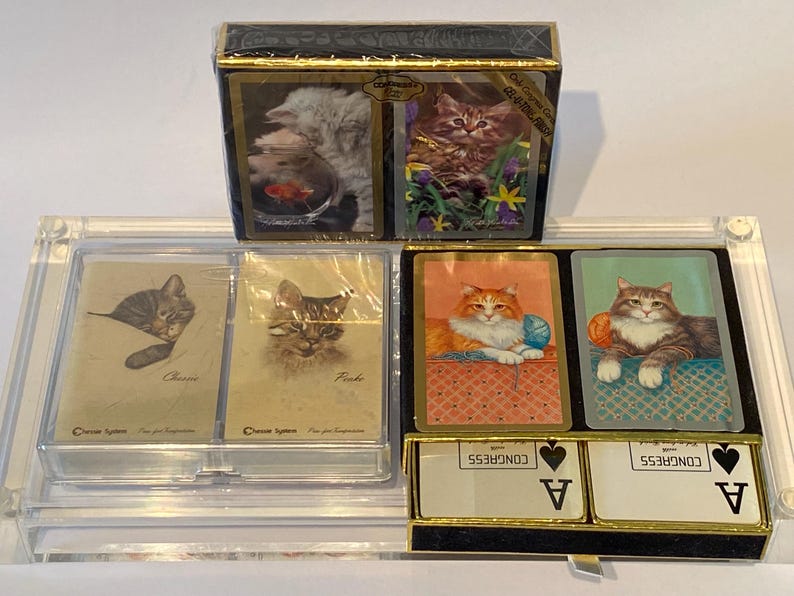 May include: A set of four playing card decks featuring different cat illustrations. The decks are in a clear plastic case with a black base. The front of the case has the text "CONGRESS" and "A" for Ace.