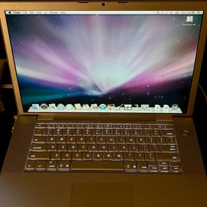 May include: A silver MacBook Pro laptop with a backlit QWERTY keyboard and a trackpad. The screen displays a purple and blue nebula wallpaper, a top menu bar with Finder, and a bottom dock containing various application icons and a Macintosh HD icon.