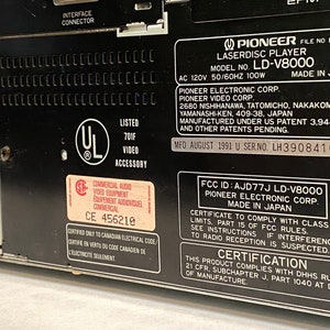 May include: Close-up of the back panel of a black Pioneer LD-V8000 Laserdisc Player. The panel includes various labels with text, including the model number, manufacturer information, and safety certifications. The unit is made in Japan.
