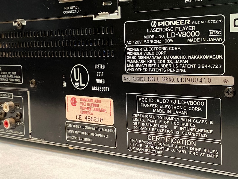 May include: Close-up of the back panel of a black Pioneer LD-V8000 Laserdisc Player. The panel includes various labels with text, including the model number, manufacturer information, and safety certifications. The unit is made in Japan.