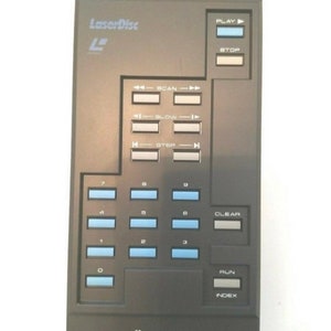 May include: A black Pioneer LaserDisc remote control unit, model AU-V6000. The remote features a series of buttons with blue and white text, including play, stop, scan, slow, step, and number keys. The LaserDisc logo is visible.