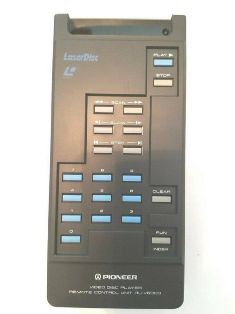 May include: A black Pioneer LaserDisc remote control unit, model AU-V6000. The remote features a series of buttons with blue and white text, including play, stop, scan, slow, step, and number keys. The LaserDisc logo is visible.