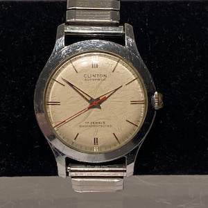 May include: A silver wristwatch with a white face and black hands. The watch face says "Clinton Automatic" and "17 Jewels Shockprotected".