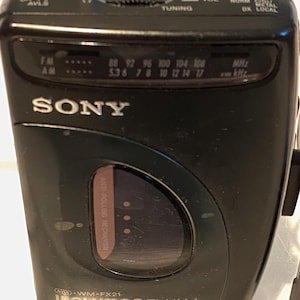 May include: Black Sony Walkman FM/AM radio with a tuning dial, volume control, and frequency display. The device has a headphone jack and the text "WALKMAN FM/AM" is visible. The radio has a built-in anti-rolling mechanism.