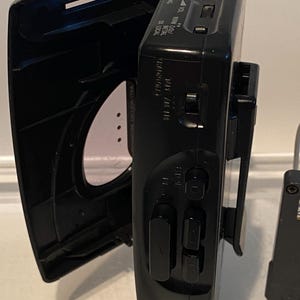 May include: Black portable cassette player with an open lid. The device has buttons for play, stop, and rewind. A white and green audio jack is inserted. The device has a clip on the back.